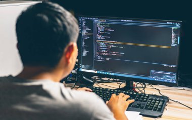 Arka planda oturan Asyalı genç adam bilgisayar programlama kullanarak çalışma istasyonunda ikili algoritma kodunu geliştirerek monitöre odaklanarak ve insanlara bulanıklaştırarak hata ayıklama yapıyor.