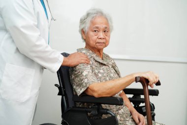 Doktor, hastanede tekerlekli sandalyede oturan Asyalı yaşlı kadın engelli hastaya yardım ediyor..  