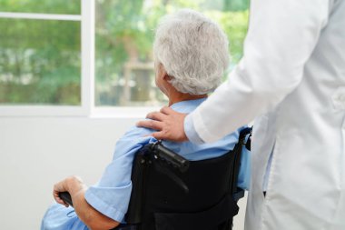 Doktor, hastanede tekerlekli sandalyede oturan Asyalı yaşlı kadın engelli hastaya yardım ediyor..