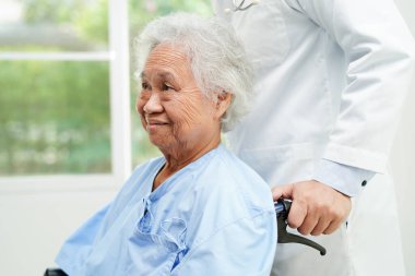 Doktor, hastanede tekerlekli sandalyede oturan Asyalı yaşlı kadın engelli hastaya yardım ediyor..