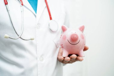 Domuzcuk kumbarası tutan Asyalı kadın doktor, tedavi masrafları veya eğitim konsepti.       