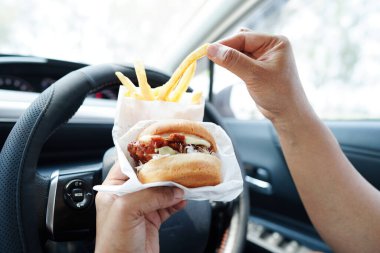 Asyalı kadın sürücü arabada hamburger ve patates kızartması yer, tehlikeli ve kaza riski taşır..