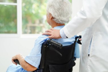 Doktor, hastanede tekerlekli sandalyede oturan Asyalı yaşlı kadın engelli hastaya yardım ediyor..