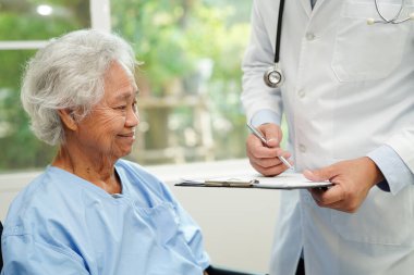 Doktor, hastanede tekerlekli sandalyede oturan Asyalı yaşlı kadın engelli hastaya yardım ediyor..