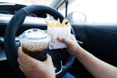 Asyalı kadın sürücü arabada patates kızartması yer ve tutar. Tehlikeli ve kaza riski taşır..