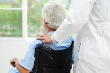 Doktor, hastanede tekerlekli sandalyede oturan Asyalı yaşlı kadın engelli hastaya yardım ediyor..