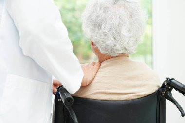 Doktor, hastanede tekerlekli sandalyede oturan Asyalı yaşlı kadın engelli hastaya yardım ediyor..