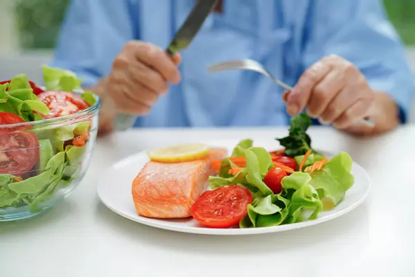 Asian elderly woman patient eating salmon stake and vegetable salad for healthy food in hospital.