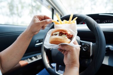 Asyalı kadın sürücü arabada hamburger ve patates kızartması yer, tehlikeli ve kaza riski taşır..