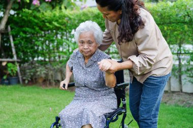 Bakıcı, parkta tekerlekli sandalyede oturan Asyalı yaşlı kadın maluliyet hastasına yardım ediyor..