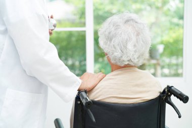 Doktor, hastanede tekerlekli sandalyede oturan Asyalı yaşlı kadın engelli hastaya yardım ediyor..