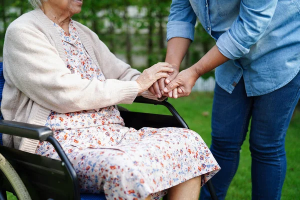 Bakıcı, parkta tekerlekli sandalyede oturan Asyalı yaşlı kadın maluliyet hastasına yardım ediyor..