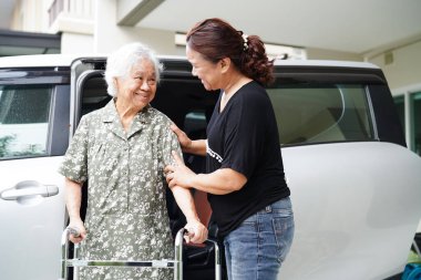 Bakıcı, Asyalı yaşlı kadın maluliyet hastasına arabasına binmesi için yardım ediyor..