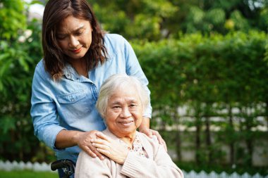 Bakıcı, parkta tekerlekli sandalyede oturan Asyalı yaşlı kadın maluliyet hastasına yardım ediyor..