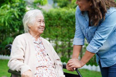 Bakıcı, parkta tekerlekli sandalyede oturan Asyalı yaşlı kadın maluliyet hastasına yardım ediyor..