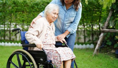 Bakıcı, parkta tekerlekli sandalyede oturan Asyalı yaşlı kadın maluliyet hastasına yardım ediyor..