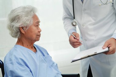 Doktor, hastanede tekerlekli sandalyede oturan Asyalı yaşlı kadın engelli hastaya yardım ediyor..