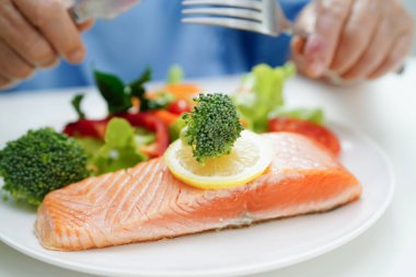 Hastanede sağlıklı yiyecekler için somon kazığı ve sebze salatası yiyen Asyalı yaşlı kadın hasta..