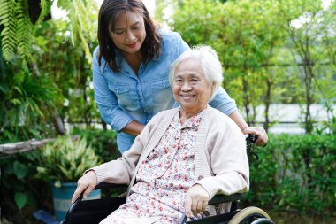 Bakıcı, parkta tekerlekli sandalyede oturan Asyalı yaşlı kadın maluliyet hastasına yardım ediyor..