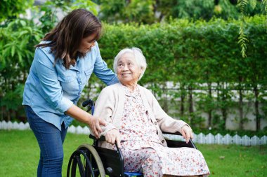 Bakıcı, parkta tekerlekli sandalyede oturan Asyalı yaşlı kadın maluliyet hastasına yardım ediyor..