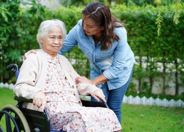 Bakıcı, parkta tekerlekli sandalyede oturan Asyalı yaşlı kadın maluliyet hastasına yardım ediyor..