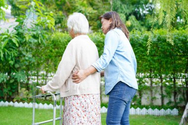 Bakıcı, Asyalı yaşlı kadın engelli hastaya parkta Walker 'la yürümesi için yardım ediyor..