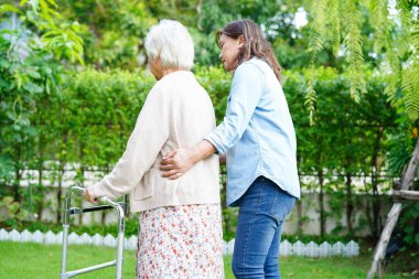 Bakıcı, Asyalı yaşlı kadın engelli hastaya parkta Walker 'la yürümesi için yardım ediyor..