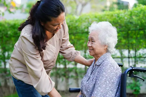 Bakıcı, parkta tekerlekli sandalyede oturan Asyalı yaşlı kadın maluliyet hastasına yardım ediyor..