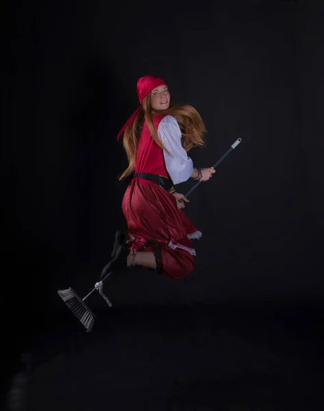 woman with long hair disguised as a pirate in a red suit and with a yellow balloon in the shape of a sword in her hand and on a black background photograph uploaded on a broomstick as a witch flying from party events