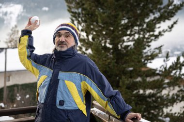 Gri saçlı, sakallı ve spor giysili gözlüklü yetişkin bir adam, bir balkonda dağın arka planına karşı duruyor, kameraya bakıyor ve bir kartopu atıyor. Yüksek kalite fotoğraf