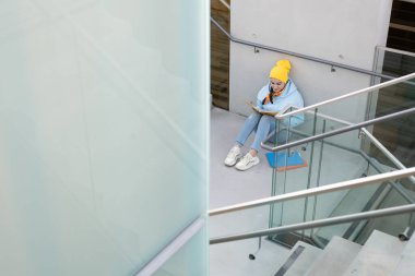 Mavi kazaklı ve sarı şapkalı bir öğrenci merdivenlerin arasında taş bir zeminde oturup kitap okuyor. Yüksek kalite fotoğraf