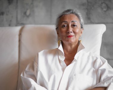 Charming lady in a loose white shirt sits on a white sofa, looks at the camera and smiles. High quality photo