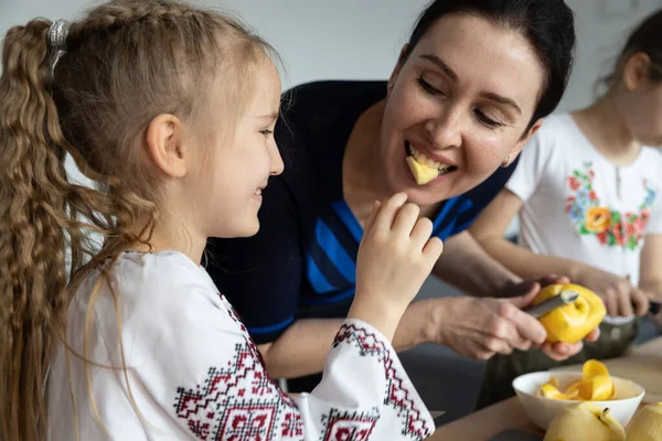 A mother and two daughters peel apples for a pie, the youngest daughter in a Ukrainian embroidered shirt treats her mother to an apple. High quality photo