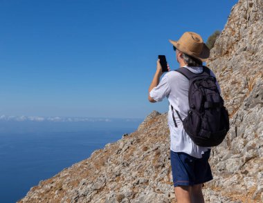 Yetişkin bir adam dik bir deniz kıyısında duruyor ve telefonuyla güzel bir Yunan manzarası olan Girit 'i çekiyor. Yüksek kalite fotoğraf