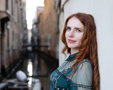 Güzel, uzun saçlı, kızıl saçlı kadın Venedik 'in su kanallarının arka planında duvara dayanıyor ve İtalya' nın kamerasına bakıyor. Yüksek kalite fotoğraf