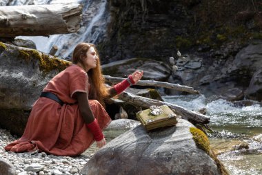Yaşlı elbiseli kızıl saçlı genç bir kadın bir dağ nehrinin kıyısında bir kayanın arka planına karşı oturur ve suya taşlar atar. Yüksek kalite fotoğraf