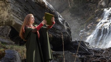 Kızıl saçlı, uzun saçlı genç bir kadın Avusturya 'daki bir şelalenin ve kayanın zemininde duran eski bir kitaba bakıyor. Yüksek kalite fotoğraf