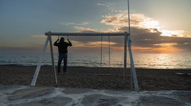 Uzun saçlı bir adam deniz kenarında salıncakta durur ve gün batımına hayran kalır. Yüksek kalite fotoğraf