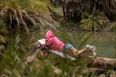 Nehir kıyısında, palmiye ağaçlarının arasında bir kız bir ağaca uzanıp kitap okuyor. Yüksek kalite fotoğraf