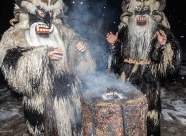 İki Noel Krampusu, karlı bir gecede Avusturya 'da sigara içilen bir ağacın yanında ellerini ısıtıyor, Gastein. Yüksek kalite fotoğraf