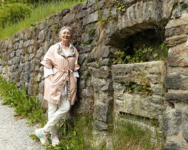 Gri saçlı bir kadın taş bir duvara yaslanmış duruyor ve kameraya gülümsüyor. Yüksek kalite fotoğraf