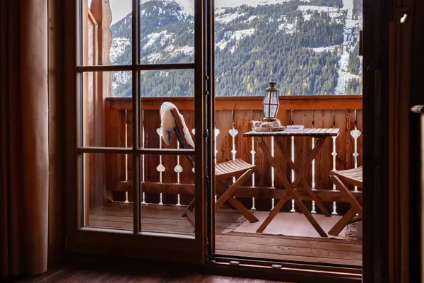 View through the balcony door to a cozy furnished balcony and a mountain slope with a ski lift and a forest area. High quality photo