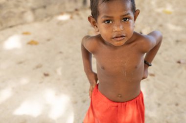 INDONEZYA, ISLAND KRI - 27 HAZİRAN 2024: Küçük bir Endonezyalı çocuk kumdaki bir ağacın gölgesinde duruyor. Yüksek kalite fotoğraf