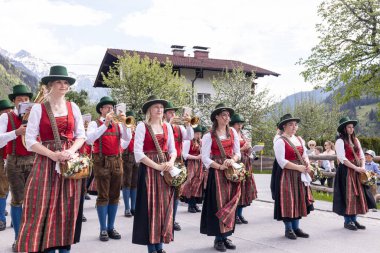 Geleneksel Avusturya kıyafeti giymiş bir grup birey bir bahar geçit törenine katılıyor. Çoğunluğu kadın olan grup üyeleri, ellerinde müzik aletleriyle sıraya girmiş durumdalar.