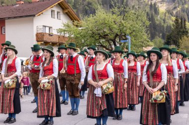 Bu resim bir grup insanı gösteriyor. Geleneksel Avusturya kıyafetleri içinde, muhtemelen bir halk grubu, bir caddede yürüyor. Renkli elbiseler ve şapkalar giyiyorlar ve ellerinde müzik aletleri var.. 