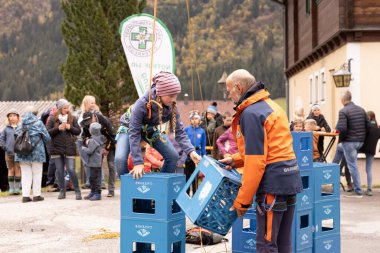 Mavi tulumlu genç bir kız, turuncu ceketli bir adam ona yardım ederken mavi plastik sandıklara tırmanıyor..