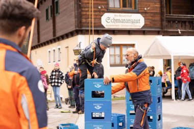 Mavi ceketli ve siyah pantolonlu genç bir çocuk emniyet kemeri takmış ve elinde bir ip tutarken mavi sandıklara tırmanıyor.. 