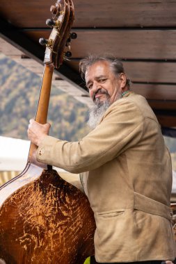 Uzun sakallı bir adam önünde bir bas gitar tutuyor. Yavaşça gülümsüyor ve doğrudan izleyiciye bakıyor.. 