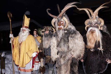 Aralarında Aziz Nicholas ve Krampus 'un da yer aldığı geleneksel kostümler giymiş bir grup insan Avusturya sokaklarında düzenlenen bir geçit törenine katılıyor. Karakterler bir çizgi üzerinde yürüyor.