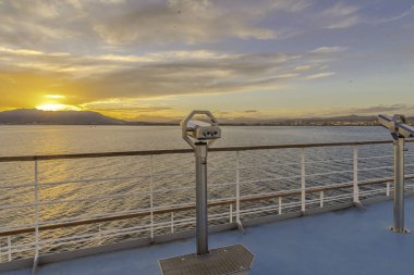 Gün batımına doğru yelken açan bir yolcu gemisinin güvertesinde dürbünler..